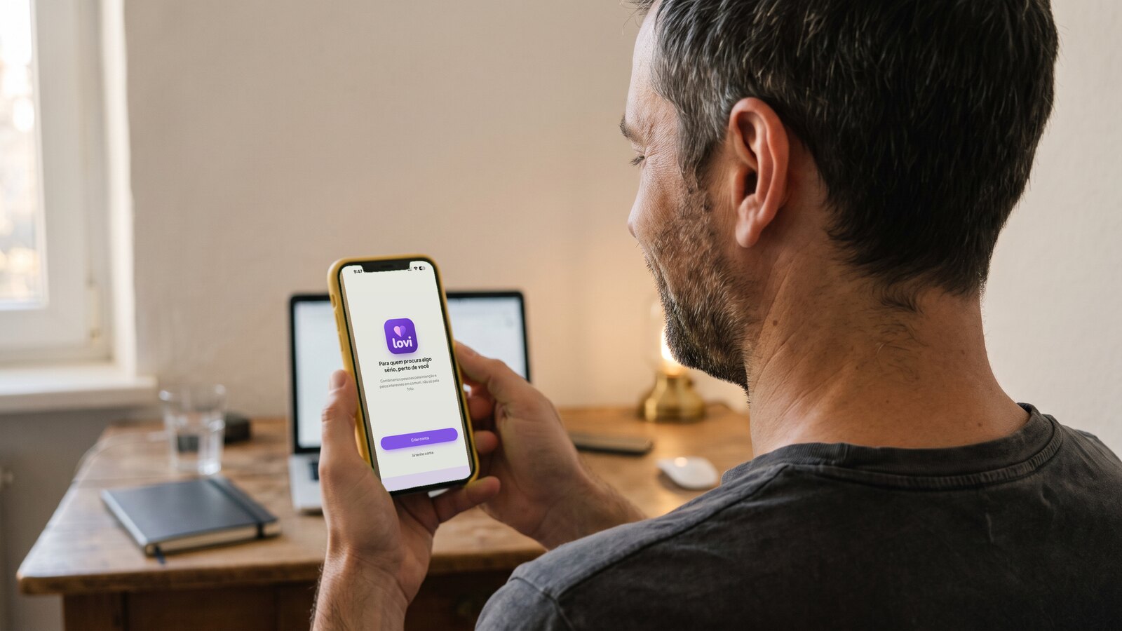 Seen over his shoulder, a man in his forties at his desk late in the day holds a phone in a plain yellow case; the screen shows the first screen of the lovi app, its logo and the line “for whoever is looking for something serious, close to you”.